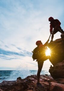 Two individuals in red jackets assisting each other to climb a steep cliff, demonstrating teamwork and support.